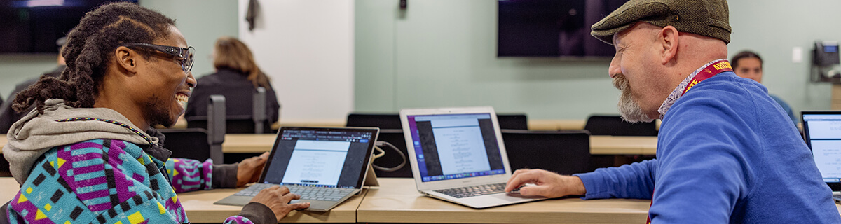 A student and a faculty member sitting at a table together looking at their laptop screens and collaborating.